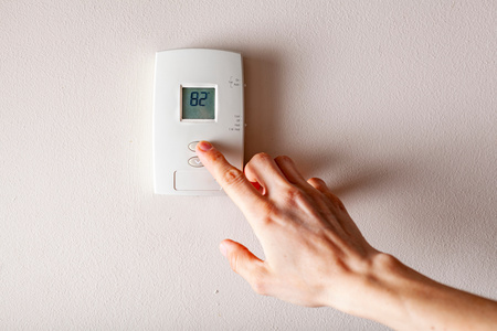 A close-up of a hand pressing the button on a digital wall thermostat displaying 82 degrees to lower the temperature.