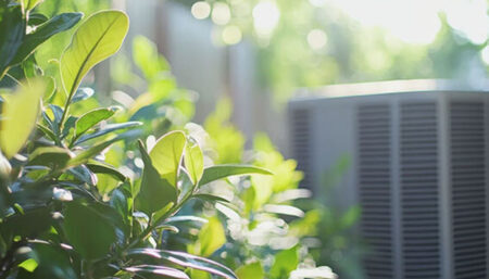 A blurred central air conditioner unit sitting behind green garden bushes during a sunny summer day.