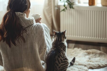 Young woman wearing a sweater freezing for winter cold with her cat. Girl is sitting over electric heater, drinking hot coffee or tea, warming up hands. Discomfort spending time at home. Coldly, chill